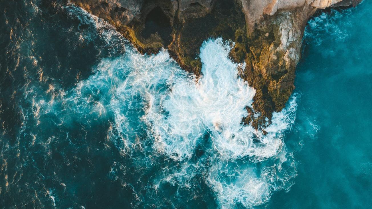 Luftfoto av bølger som treffer steinete klipper, med hvitt skum og turkist hav rundt.
